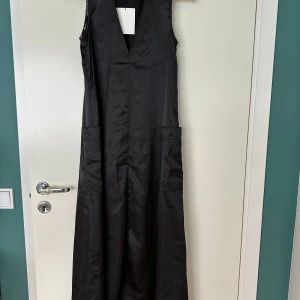 Svart långklänning från Totême - Svart ärmlös långklänning från Totême med djup v-ringning och två stora fickor framtill. Klänningen har subtila mönstrade detaljer längs kanterna och nedtill, vilket ger en cool kontrast. Materialet är mjukt och har ett lätt glansigt utseende.