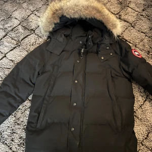Svart dunjacka från Canada Goose - Säljer en svart dunjacka från Canada Goose med fet pälskant på huvan och klassisk logga på ärmen. Jackan har dragkedja och knappar framtill, stora fickor och är perfekt för kalla vinterdagar. Modellen är lång och har en riktigt varm vibe.