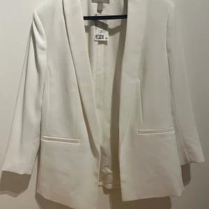 Vit kavaj från H&M, storlek 38 - Snygg vit kavaj från H&M i storlek 38. Klassisk rak modell med smala slag och två fickor framtill. Stilren och minimalistisk design, perfekt för att lyfta vilken outfit som helst. Tillverkad i ett mjukt och lätt material som känns bekvämt hela dagen.