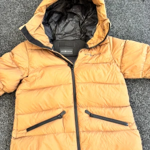 Ser gul ut men den är nogatfärgad - Orange dunjacka från Sail Racing med huva och svarta detaljer. Jackan har två snedställda fickor med dragkedja och quiltad design. Perfekt för kyliga dagar och har en sportig vibe.