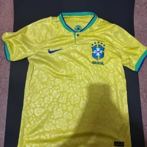 Brazil Nike fotbollströja - Gul Brasilien fotbollströja från Nike med blått Swoosh, gröna och blå detaljer på krage och ärmslut. CBF-emblem på bröstet och subtilt mönster i tyget. Tillverkad i Dri-FIT-material som andas och känns lätt mot kroppen.