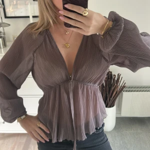 Plisserad brun blus från Zara - Säljer en snygg plisserad blus från Zara i en mjuk brun nyans. Blusen har djup v-ringning, långa puffärmar och ett knytband under bysten som ger en feminin siluett. Materialet är lätt och luftigt, perfekt för att skapa en cool och trendig look.