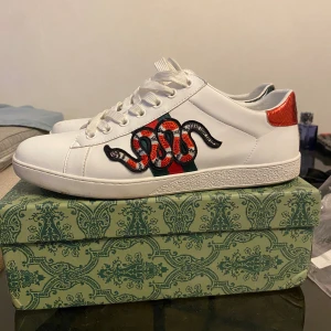 Gucci sneakers med ormmotiv - Säljer ett par vita Gucci sneakers med broderad röd och svart orm på sidan, klassiska gröna och röda ränder samt röd och grön detalj på hälen. Skorna har snörning och platt sula, tillverkade i skinn med lyxig känsla. Kommer med originalkartong.
