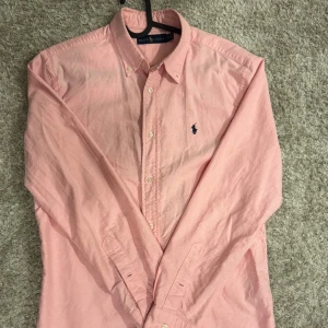 Rosa skjorta från Ralph Lauren S - Snygg rosa skjorta från Ralph Lauren i storlek S. Klassisk button-down krage, långärmad och med det ikoniska blå broderade logot på bröstet. Skjortan är tillverkad i mjuk bomull och har knappar framtill. Perfekt för dig som gillar stilrena och tidlösa plagg.