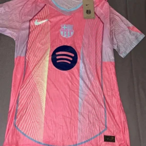 Rosa FC Barcelona fotbollströja Nike - Säljer en snygg FC Barcelona fotbollströja från Nike i rosa med ljusblå och gula detaljer. Tröjan har korta ärmar, klubbmärke på bröstet och Spotify-logga i mörkblått. Materialet är lätt och andas, perfekt för match eller träning.