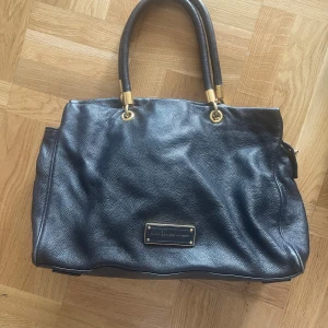 Svart handväska från Marc by Marc Jacobs - Snygg svart handväska från Marc by Marc Jacobs i äkta skinn med guldfärgade metalldetaljer. Väskan har två stadiga handtag, dekorativ logoplatta framtill och dragkedja med guldfärgad zip. Perfekt storlek för att få plats med allt du behöver.