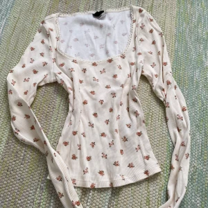 Blommig ribbad topp från Divided XS - Supersöt ribbad topp från Divided i ljusbeige med små blommor i rosa och grönt. Toppen har lång ärm, fyrkantig ringning med spetskant och figurnära passform. Perfekt för dig som gillar romantisk och feminin stil.