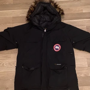Svart parkas från Canada Goose - Svart parkasjacka från Canada Goose med stor huva och fuskpäls, flera praktiska fickor och klassisk logga på bröstet. Jackan har dragkedja framtill och är perfekt för kalla vinterdagar. Kom privat för fler bilder 