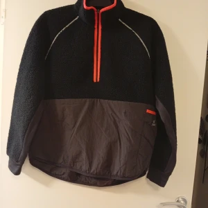 Svart fleece-tröja från SOC med half zip - Svart och mörkgrå fleece-tröja från SOC med orange detaljer och half zip framtill. Tröjan har lång ärm, hög krage och reflekterande detaljer. Perfekt för kyliga dagar och träning utomhus.