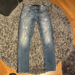 Replay ljusblå slitna jeans - Säljer ett par ljusblå jeans från Replay med snygga slitningar och tvättade detaljer. Klassisk femficksmodell med raka ben och läderpatch bak i midjan. Jeansen har normal passform och är tillverkade i mjukt denimtyg för en skön känsla.