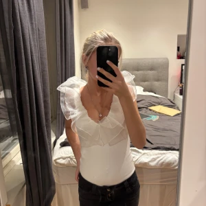 Vit volang body från Amisu XS - Säljer en vit body från Amisu i storlek XS med transparenta volanger runt axlar och halsringning. Toppen är ärmlös och har en slim passform med ribbad struktur. Perfekt för dig som gillar statement-detaljer och en feminin look. Säljer då den inte används.