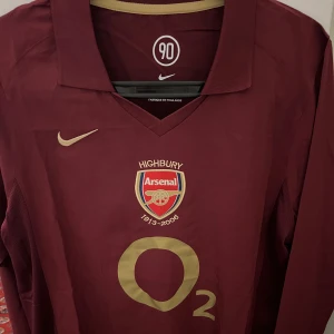 Arsenal Highbury 2005/06 fotbollströja Nike - Säljer en ikonisk Arsenal Highbury 1913-2006 fotbollströja från Nike. Tröjan är vinröd med guldfärgade detaljer, klubbmärke och O2-logga på bröstet. Klassisk krage och långärmad modell i lätt och ventilerande material. Perfekt för samlare eller Arsenal-fans, priset går att diskutera vid snabb affär 