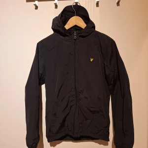 Svart vindjacka från Lyle & Scott - Svart vindjacka från Lyle & Scott med huva och dragkedja framtill. Jackan har två fickor med knappstängning och den klassiska gula loggan på bröstet. Tillverkad i lätt polyester, perfekt för blåsiga dagar. Storlek Junior 14/15 år 