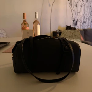 Svart axelväska med dragkedja - Snygg svart axelväska i stilren från Ur&pen original pris var 400-500kr, avlång form med justerbar rem och praktisk dragkedja upptill. Perfekt storlek för att få plats med det viktigaste och passar till många olika outfits. Materialet är slätt och ger en clean vibe.
