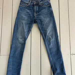 Blå jeans från Nudie Jeans, modell steady Eddie ll - Säljer ett par klassiska blå jeans från Nudie Jeans, modell steady Eddie. Jeansen har en slim passform med raka ben och snygga slitningar framtill. Orangea sömmar och femficksdesign. Perfekta för dig som gillar en stilren och tidlös look med snygg tvätt.