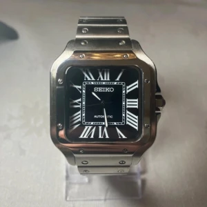 Seiko Santos - Snygg Seiko mod klocka med fyrkantig boett i borstat stål och svart urtavla med vita romerska siffror. Länken är i stål och har ett robust viklås. Genomskinlig baksida visar det mekaniska verket. Perfekt för dig som gillar stilren design.