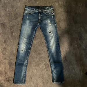 Replay blå jeans med slitningar, stl 32 - Snygga blå jeans från Replay med slitna detaljer och cool tvätt. Klassisk femficksmodell med Replay-logga på fickan och läderpatch bak. Smal passform och normal midja, perfekt för en avslappnad streetstil.