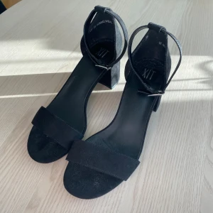 Svarta sandaler med klack från XIT - Svarta sandaler från XIT med bred rem över tårna och justerbar ankelrem med spänne. Snygg blockklack och rund tå ger en stilren look. Materialet är mocka och sulan är platt framtill.