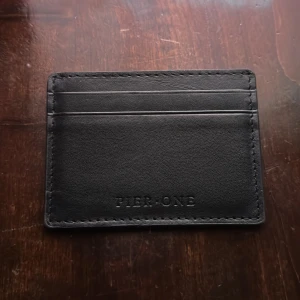 Svart kortfodral från Pier One - Slimmat svart kortfodral från Pier One i stilren design med präglad logga. Fodralet har flera fack för kort och är tillverkat i mjukt skinn med snygga sömmar runt kanterna. Perfekt för dig som vill ha ett minimalistiskt och praktiskt sätt att bära dina kort.