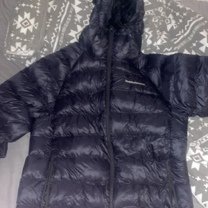 Svart dunjacka Peak Performance L - Säljer en svart dunjacka från Peak Performance i storlek L. Jackan har huva, hel dragkedja och klassisk quiltad design. Perfekt för kalla dagar och har en stilren look med broderad logga på bröstet.