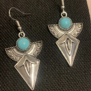 Silvriga örhängen med turkos detalj - Unika örhängen i silverfärgad metall med geometrisk form och en rund turkos sten upptill. Halvcirkelformad dekoration med små prickar och en spetsig, graverad diamantformad del. Perfekta för dig som gillar boho eller statement-smycken.