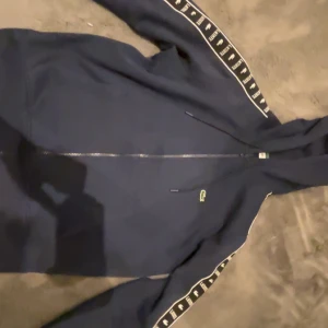 Mörkblå zip-hoodie från Lacoste M - Mörkblå hoodie från Lacoste med dragkedja och huva. Klassisk krokodillogga på bröstet och vita Lacoste-band längs ärmarna. Tillverkad i 80% bomull och 20% polyester. Perfekt för en sportig och avslappnad stil.