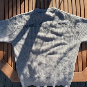 Grå stickad tröja Comme des Garçons - Riktigt snygg grå stickad tröja från Comme des Garçons Homme. Tröjan har ribbad struktur, rund hals och tryckt logga på ryggen. Perfekt för dig som gillar stilrena och exklusiva plagg med streetvibe.
