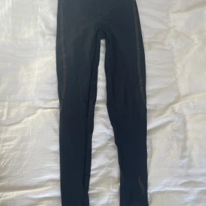 Svarta träningstights - Svarta träningstights från Casall Sculpture med glansiga detaljer längs benen och logga på vaden. Tight passform och snörning i midjan. Är i stretchigt syntetmaterial.