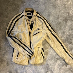 Beige skinnjacka med svarta ränder  - Snygg beige skinnjacka från som köptes second hand med svarta ränder längs ärmarna. Jackan har hög krage med spänne, flera dragkedjor framtill och en cool bikerstil. .