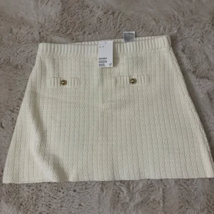 Vit kortkjol från H&M i bouclé - Stilren vit kortkjol från H&M i bouclé-liknande material. Kjolen har ribbad midja och två dekorativa fickor framtill med guldknappar. Perfekt för dig som gillar en klassisk och chic look.