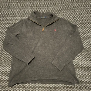 Ralph Lauren half zip tröja  - Mörkgrå långärmad tröja från Polo Ralph Lauren med klassisk röd broderad logga på bröstet. Tröjan har en snygg half zip-dragkedja och ribbade muddar. Perfekt för lager-på-lager och chill stil.