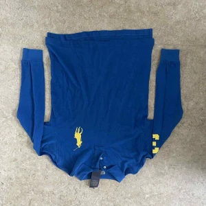 Blå långärmad pikétröja Ralph Lauren - Blå långärmad pikétröja från Polo Ralph Lauren med gul broderad logga på bröstet och gula siffror på ena ärmen. Tröjan har klassisk krage, två knappar och är i custom fit. Tillverkad i mjuk bomull, perfekt för en clean och sportig look.