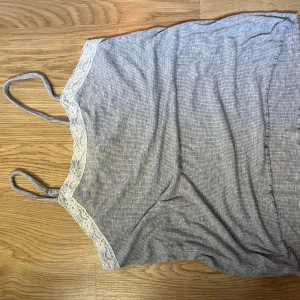 Grå ribbad linnetopp med spets - Säljer en ljusgrå ribbad linnetopp med tunna axelband och vit spetskant längs urringningen. Toppen har en enkel och stilren look med diskreta ränder och är perfekt för varma dagar.