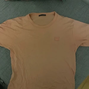 Ljusrosa t-shirt från Acne Studios - Säljer en ljusrosa t-shirt från Acne Studios med klassisk rund hals och det ikoniska Face-logotypen broderad på bröstet. T-shirten är i mjuk bomull och har en clean, enkel look som passar perfekt till jeans eller shorts. Fint skick, storlek xs