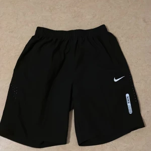 Svarta träningsshorts från Nike - Säljer ett par svarta träningsshorts från Nike med elastisk midja och vita logodetaljer. Shortsen har praktiska fickor och är tillverkade i ett lätt och snabbtorkande material, perfekta för intensiva träningspass eller löpning.