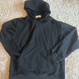 Svart hoodie från Stone Island - Svart hoodie från Stone Island med klassisk logotyp-patch på ärmen. Tröjan har huva, magficka och ribbade muddar. 