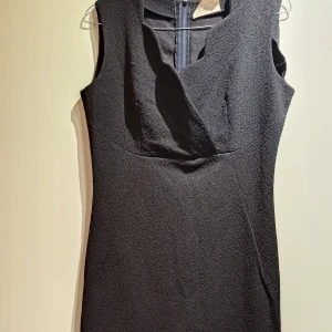 Svart ärmlös klänning från Labelle. Vintage - Snygg svart ärmlös vintage klänning från Labelle i crimplene-material. Klänningen har en figurnära passform, v-ringad omlottliknande urringning och dragkedja i ryggen. Tyget har en diskret struktur som ger extra karaktär. Perfekt för dig som gillar klassisk och stilren look. Fler bilder kan skickas vid intresse.