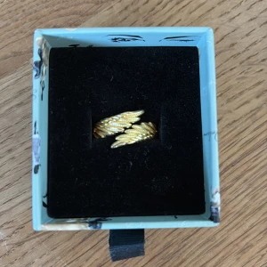 Guldig ring med fjäderdetaljer - Säljer en snygg guldig ring med två fjäderformade detaljer som möts framtill. Ringen har en cool och unik design som verkligen sticker ut!! Vet inte vart den är ifrån 💗💕