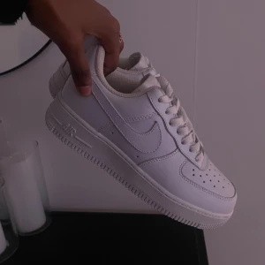 Nike Air Force 1 Vita - Fräscha Nike Air Force 1 sneakers i helvitt med klassisk låg siluett. Skorna har snörning, perforerad tå och platt sula. Tillverkade i skinn med ikonisk Swoosh på sidan. Perfekta för dig som gillar clean och stilren streetwear.