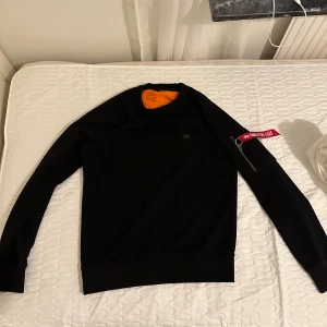 Svart Alpha Industries sweatshirt - Svart sweatshirt från Alpha Industries med orange insida och klassisk patch på bröstet. Ärmen har en cool ficka med dragkedja och röd 'Remove Before Flight'-tag. Perfekt för dig som gillar streetwear och detaljer som sticker ut.