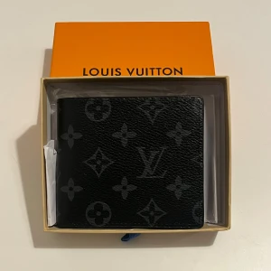 Svart plånbok med monogram - Snygg svart plånbok med klassiskt monogrammönster. Plånboken har flera kortfack, fönster för ID och är tillverkad i exklusivt skinn. Perfekt storlek för fickan och levereras i originalask.