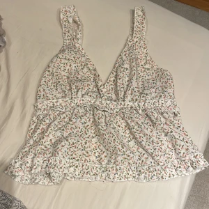 Blommig vit linnetopp med volang - Supersöt vit linnetopp med små blommor i rosa och grönt. Toppen har breda axelband, v-ringning och volangdetaljer längs midjan och nederkanten. Perfekt för varma dagar och ger en romantisk vibe.