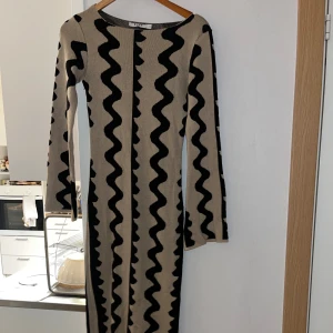 Beige och svart långklänning från NA-KD - Lång stickad klänning från NA-KD i beige med svarta vågiga ränder. Klänningen har lång ärm, rund halsringning och ett coolt grafiskt mönster som verkligen sticker ut. Perfekt för dig som gillar unika plagg med skön passform. Formar kroppen på ett galet sätt 🖤