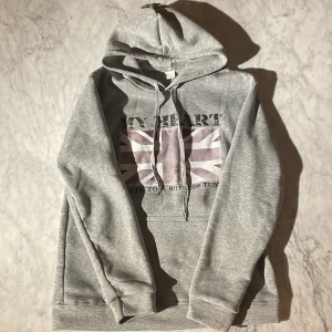 Grå hoodie med brittisk flagga - Säljer en grå hoodie med tryck av den brittiska flaggan och texten 'MY HEART BEATS TO BRITISH TUNES' på bröstet. Tröjan har huva med snörning och en klassisk känguruficka framtill. Perfekt för dig som gillar streetstyle och vill ha en chill look.