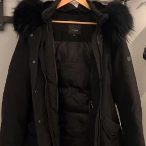 Svart parkas från Rockandblue strl 44 - Säljer en svart parkas från Rockandblue med stor huva och fluffig svart päls. Jackan har dragkedja, knappar och flera fickor framtill. Perfekt för kalla dagar med sin vadderade insida och snygga detaljer. Storlek 44.