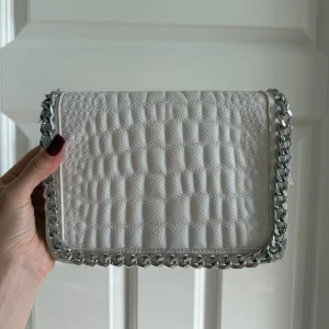 Vit quiltad axelväska med kedja - Snygg vit axelväska med quiltad struktur och grov silverfärgad kedja runt kanten. Väskan har flera fack och dragkedja på insidan, perfekt för att hålla ordning på dina grejer. Materialet är skinnimitation och ger en lyxig känsla.