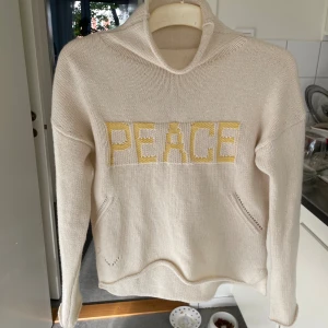 Beige stickad hoodie med PEACE-tryck - Beige stickad hoodie med broderad text 'PEACE' i gult framtill. Tröjan har lång ärm, känguruficka och en mysig huva. Perfekt för dig som gillar chill vibes och vill ha något unikt i garderoben.