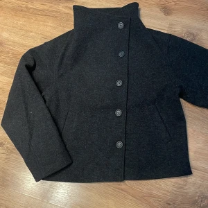 Svart kappa med hög krage och knappar - Stilren svart kappa med hög krage och raka ärmar. Jackan har fem knappar framtill och två snedställda fickor. Perfekt för dig som gillar minimalistisk och clean look. Ändast använd 1-2 gånger, nypris 400🤍