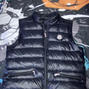 Moncler väst - Snygg svart dunväst från Moncler med glansig finish och klassisk logga på bröstet. Västen har ståkrage, två dragkedjeförsedda fickor och quiltad design. Perfekt för lager-på-lager och streetstyle. Dragkedja framtill och coola detaljer på insidan. Pris kan diskuteras privat