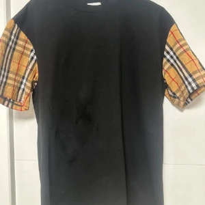 Burberry svart t-shirt med rutiga ärmar - Svart t-shirt från Burberry med klassiska rutiga kortärmar i beige, rött och svart. Snygg kontrast mellan den enfärgade kroppen och de ikoniska ärmarna. Rund halsringning och mjukt bomullsmaterial. Perfekt för dig som gillar stilrena designer med en twist.